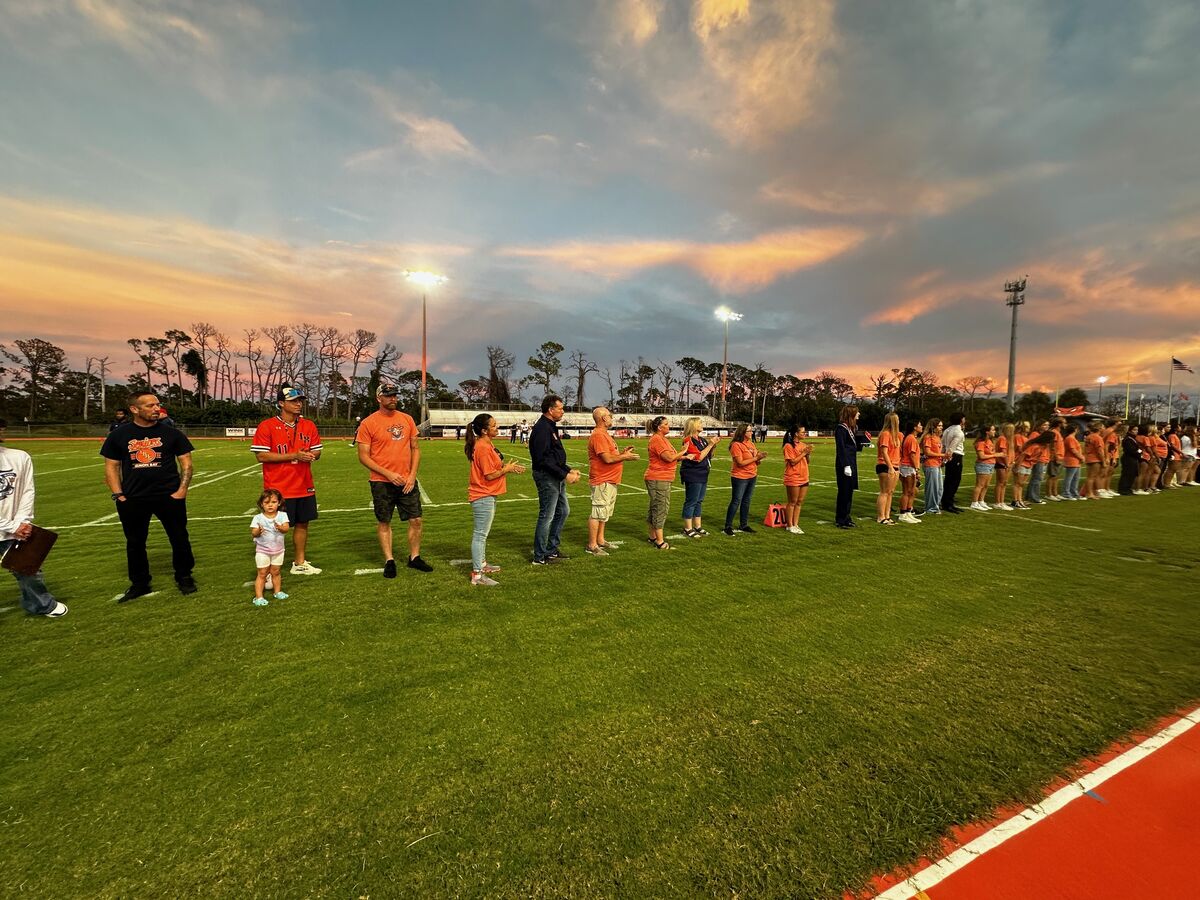 A group of people stand on a football field at dusk, under a colorful sky.