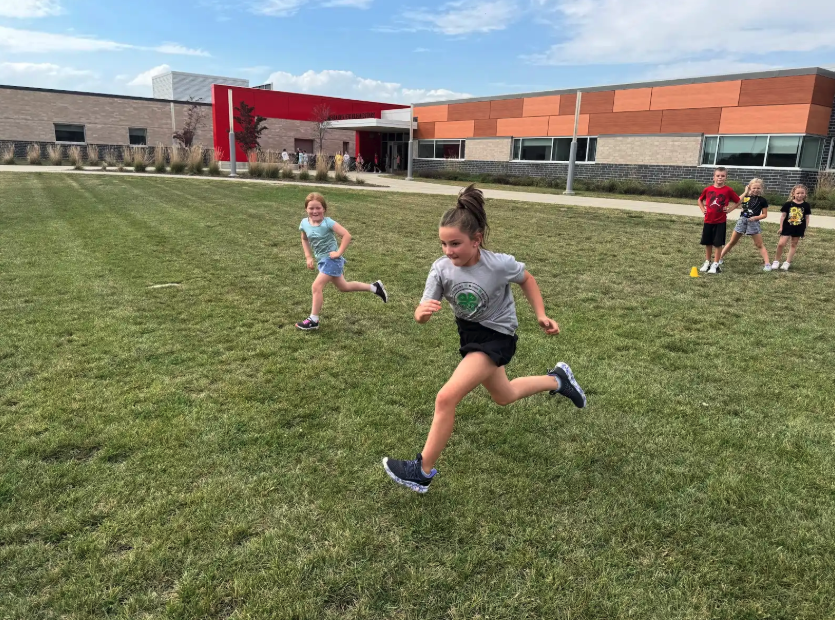 Children run across a grassy field in front of a school building.