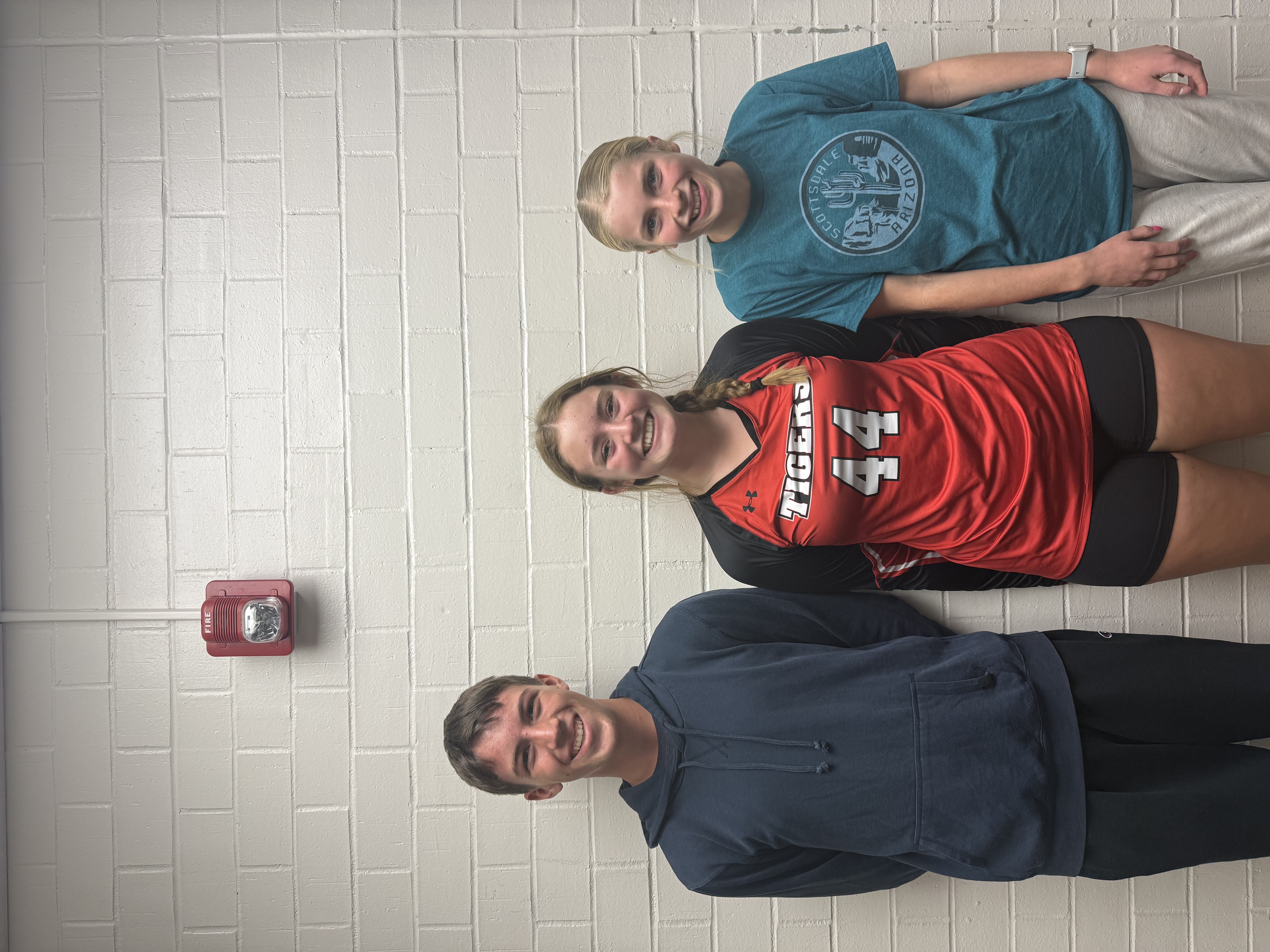Three smiling young people stand in front of a white brick wall.