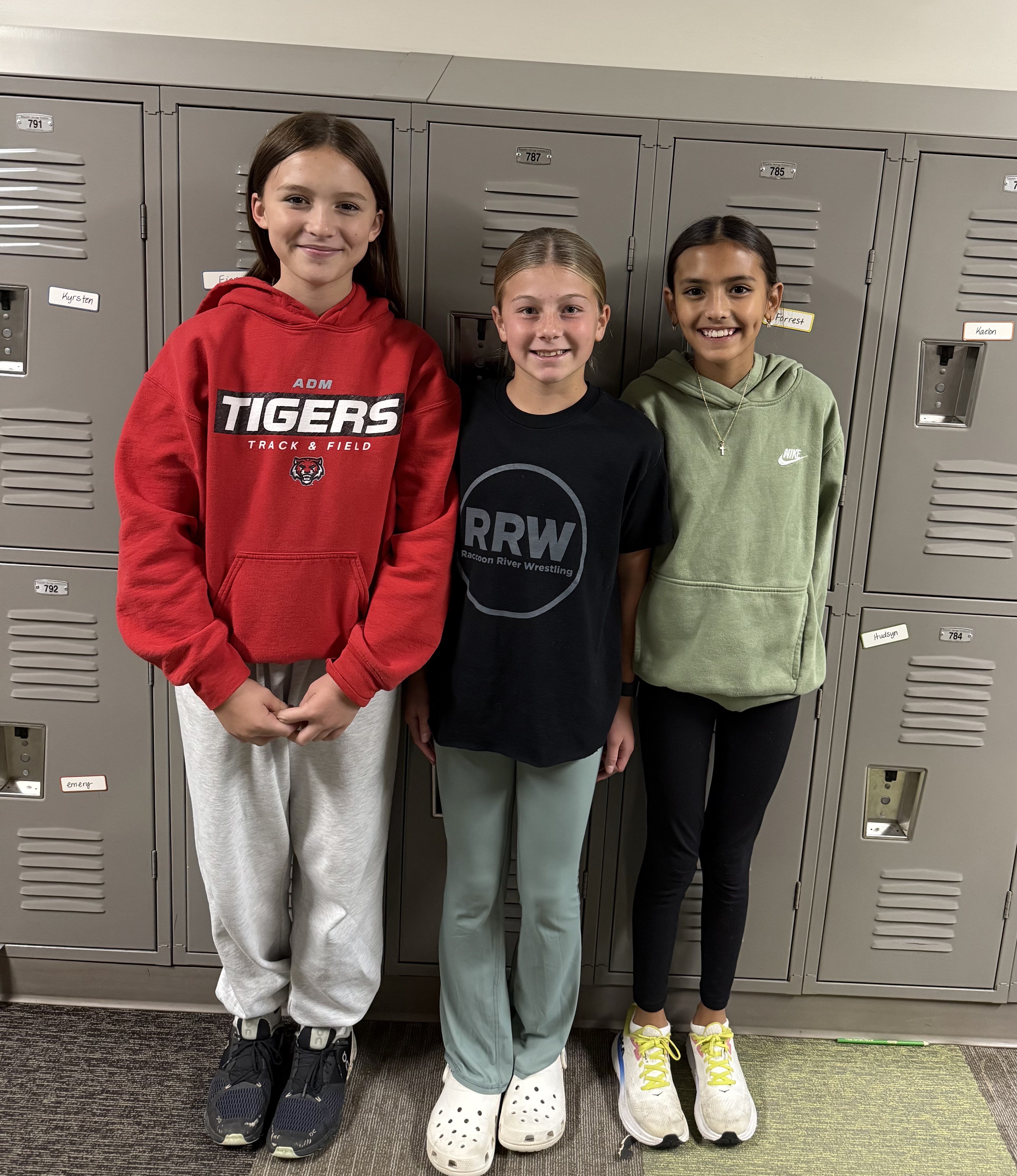 Three young girls stand together in front of school lockers, smiling at the camera.