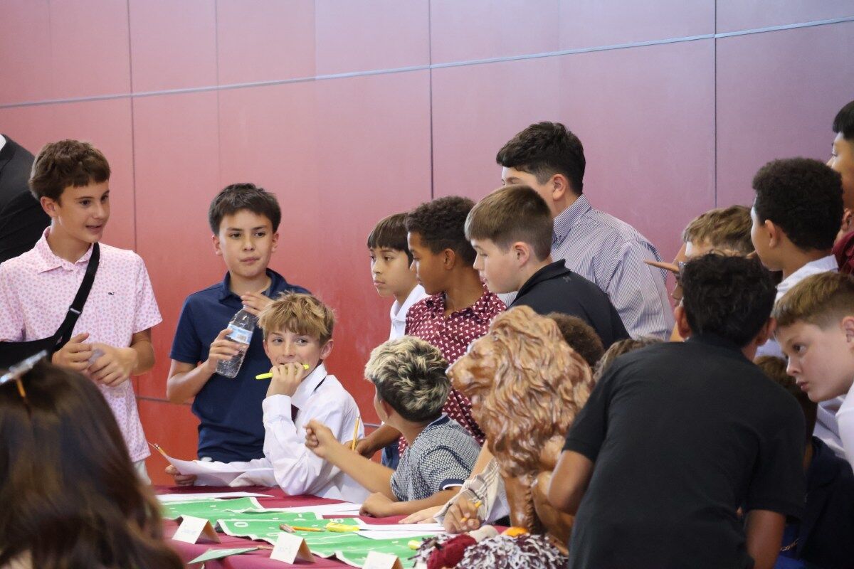 A group of boys gather around a table, some holding pencils and papers.