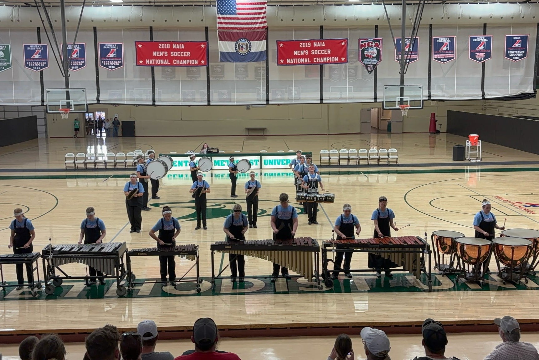 Many CHS drummers wearing light blue tops and bandana headbands, are situated on a gym floor with their percussion instruments, appearing to play