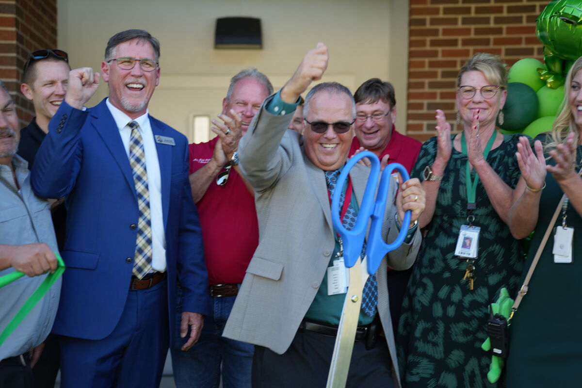 A group of people celebrate a ribbon-cutting ceremony with large blue scissors.