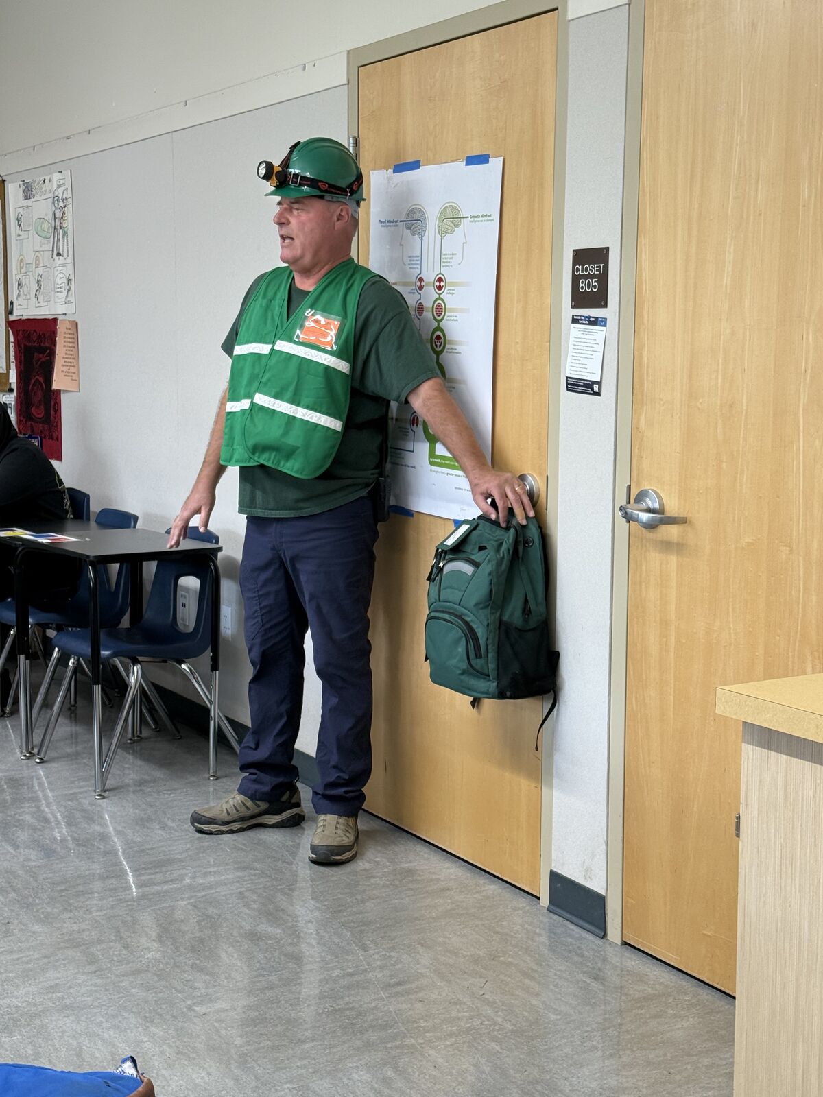 A man in a green vest and hard hat stands in a classroom, holding a backpack.