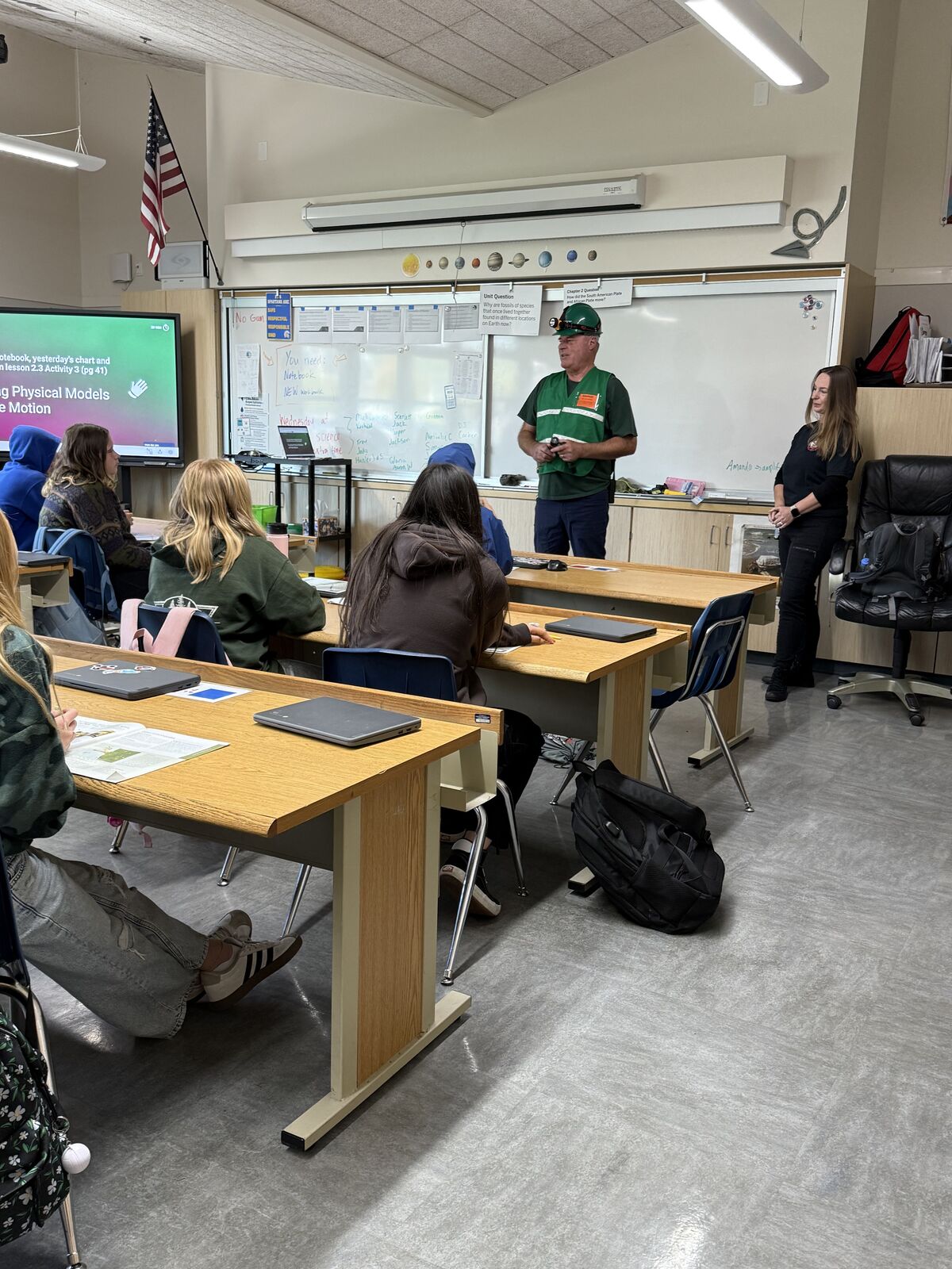 A classroom scene with students listening to a presentation by a man in a hard hat.