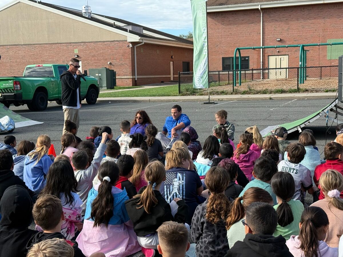 A group of children sit on the ground, listening to adults speak outdoors.