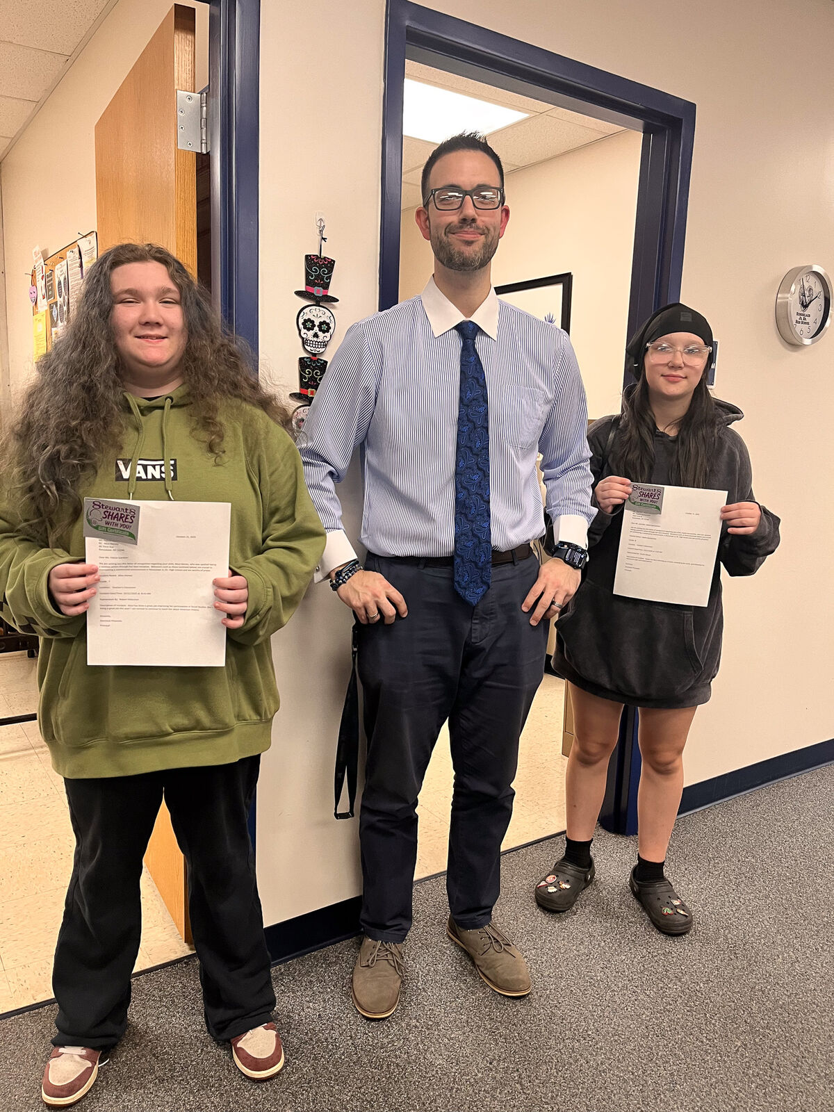 A teacher stands with two students, all holding papers, in a school hallway.