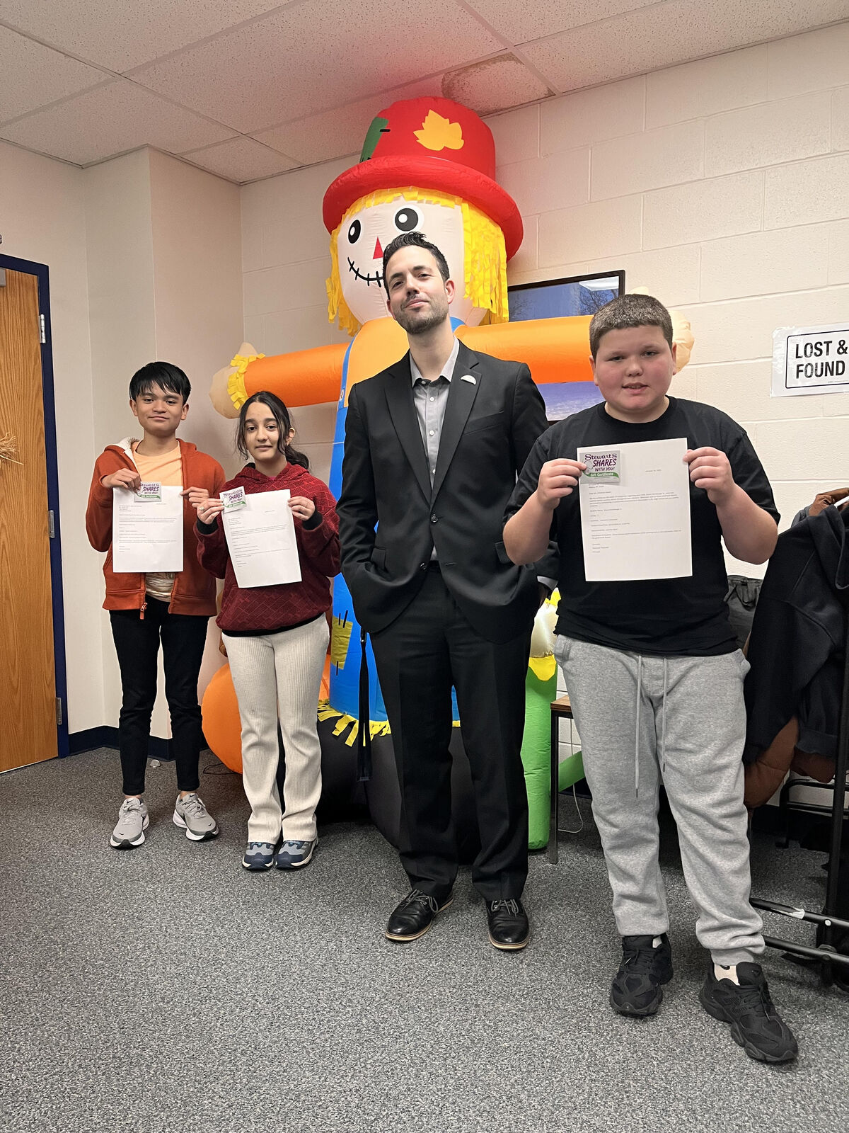Four people stand in a hallway, holding papers, with a large inflatable scarecrow behind them.