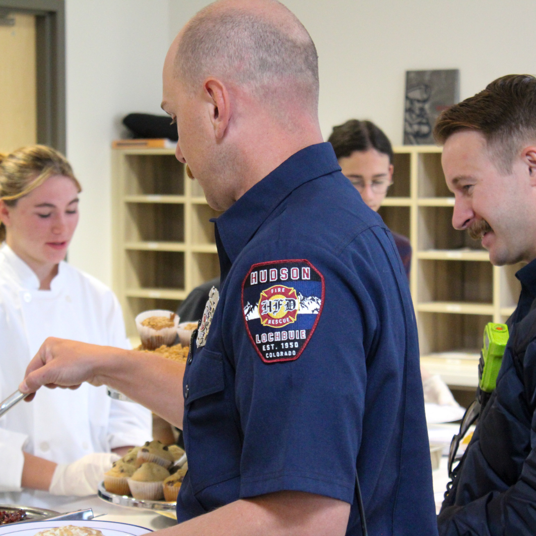 A firefighter selects a muffin from a tray, assisted by a student
