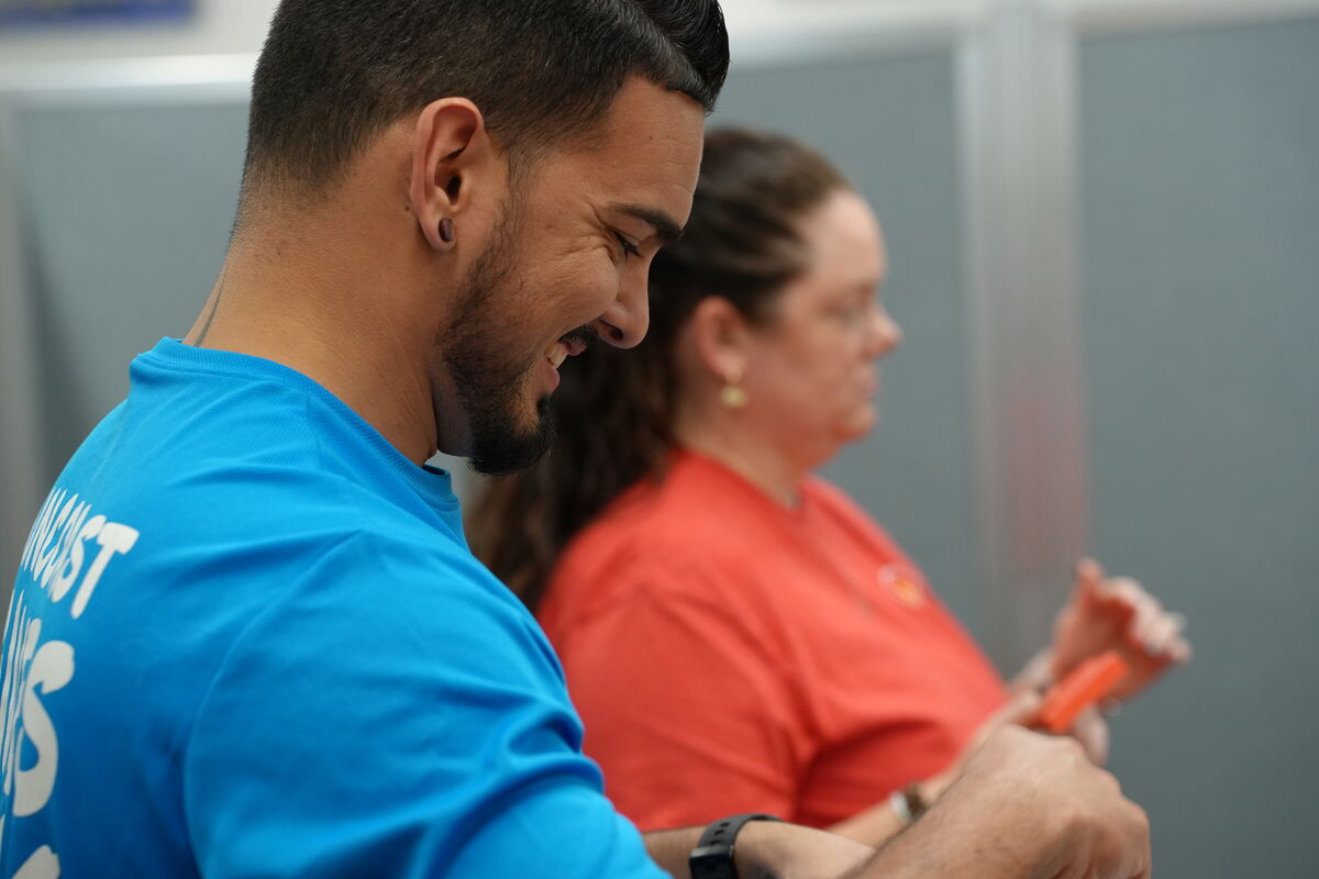 A man in a blue shirt smiles, looking down, with another person in the background.