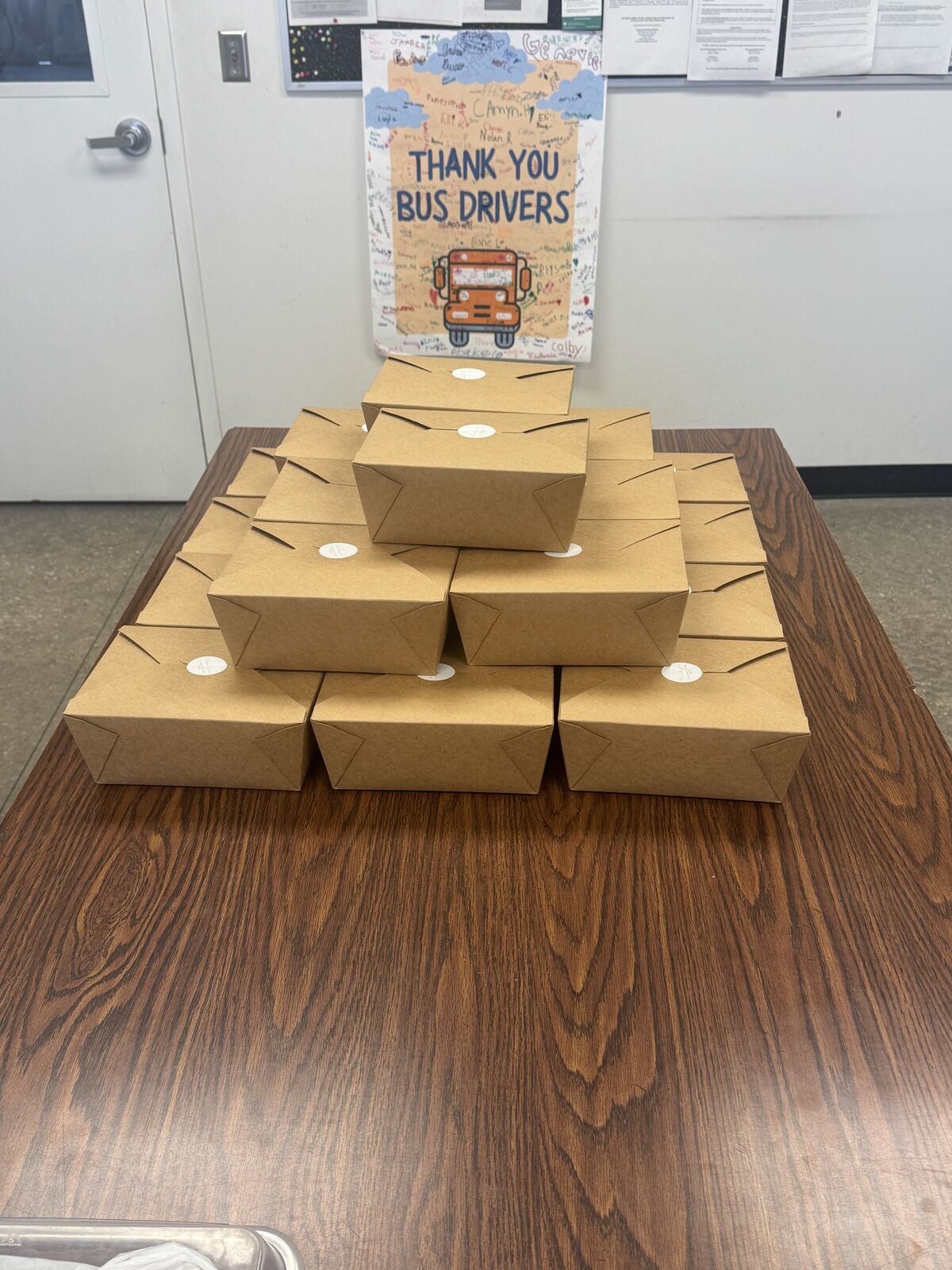 A stack of boxed meals sits on a wooden table, with a 'Thank You Bus Drivers' sign in the background.