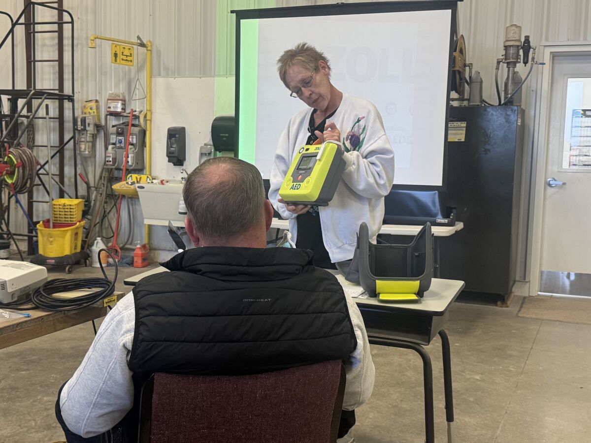 A woman demonstrates an AED to a seated man in a workshop setting.