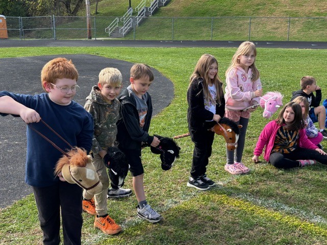Children stand on a grassy field, holding stick horses.