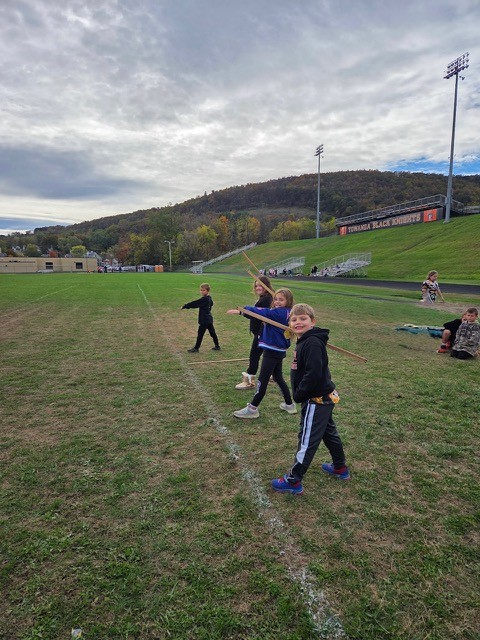 Children practice with wooden sticks on a grassy field under a cloudy sky.