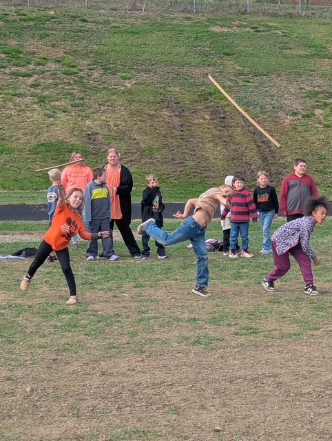 Children play a game on a grassy field, with a wooden stick in the air.