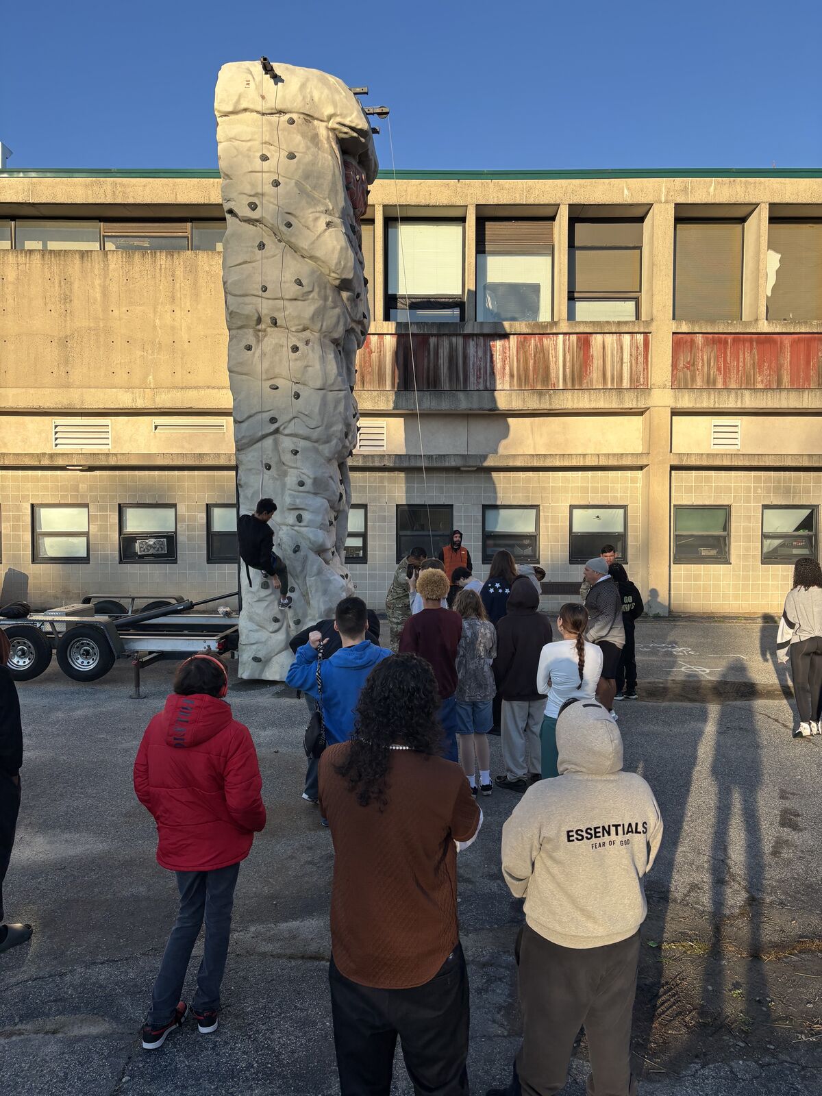 A group of people stand near a climbing wall, with a building in the background.