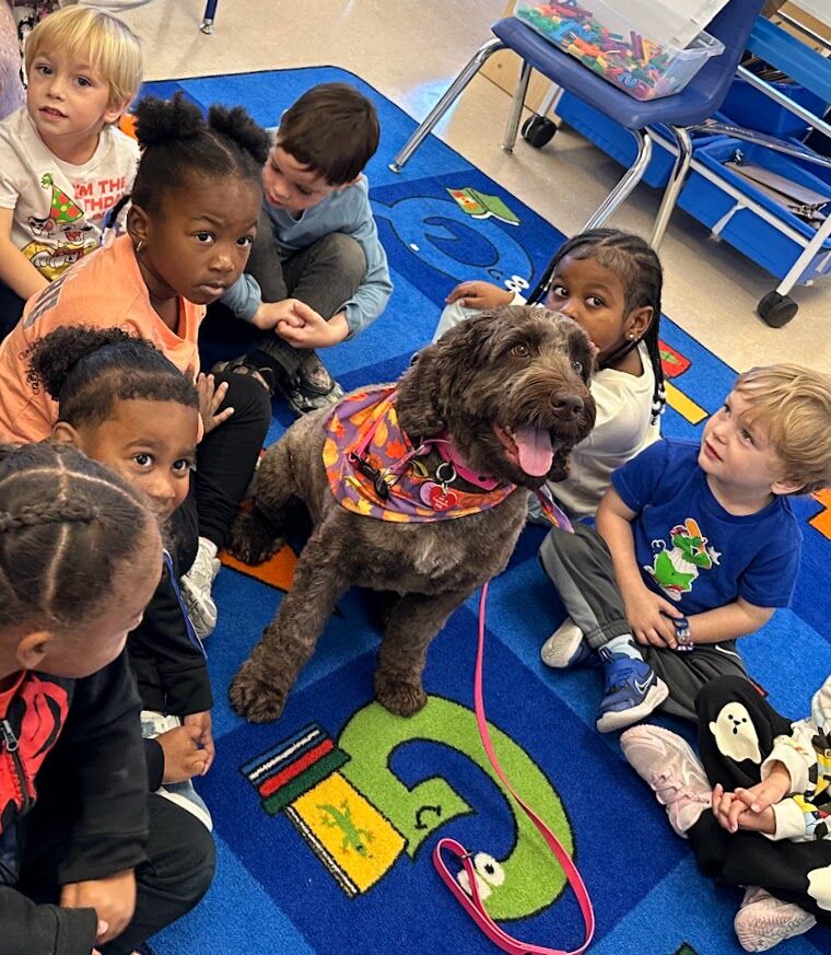 A group of children gather around a friendly dog in a classroom setting.