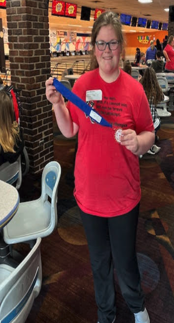 A young woman smiles, holding a medal and ribbon in a bowling alley.