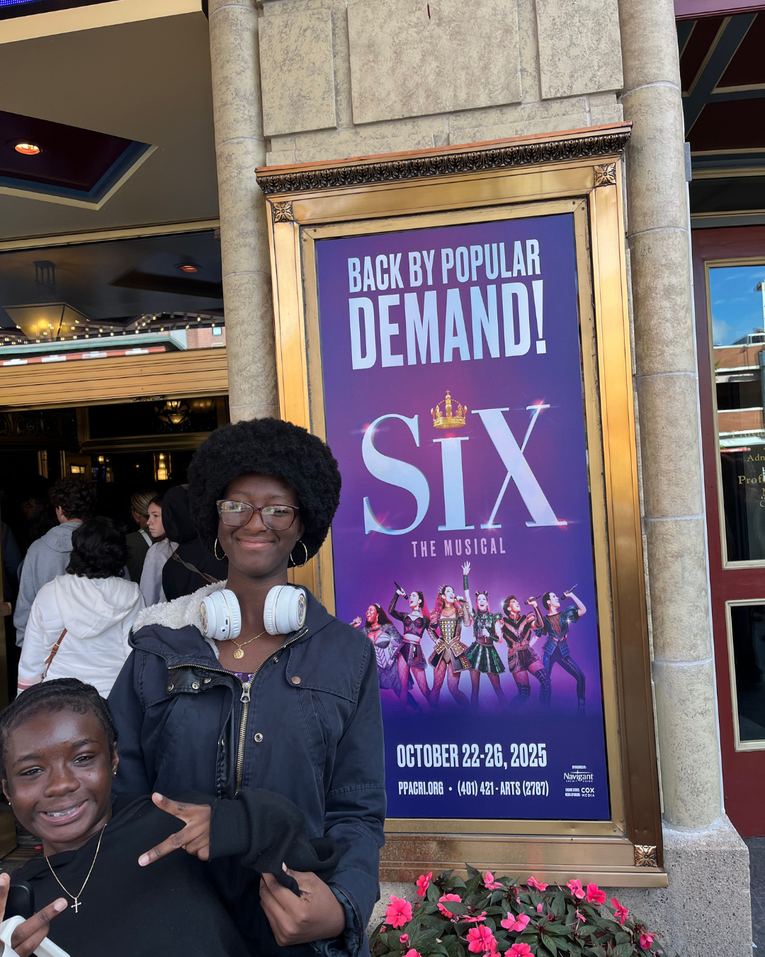 Two young women pose in front of a 'Six the Musical' poster outside a theater.