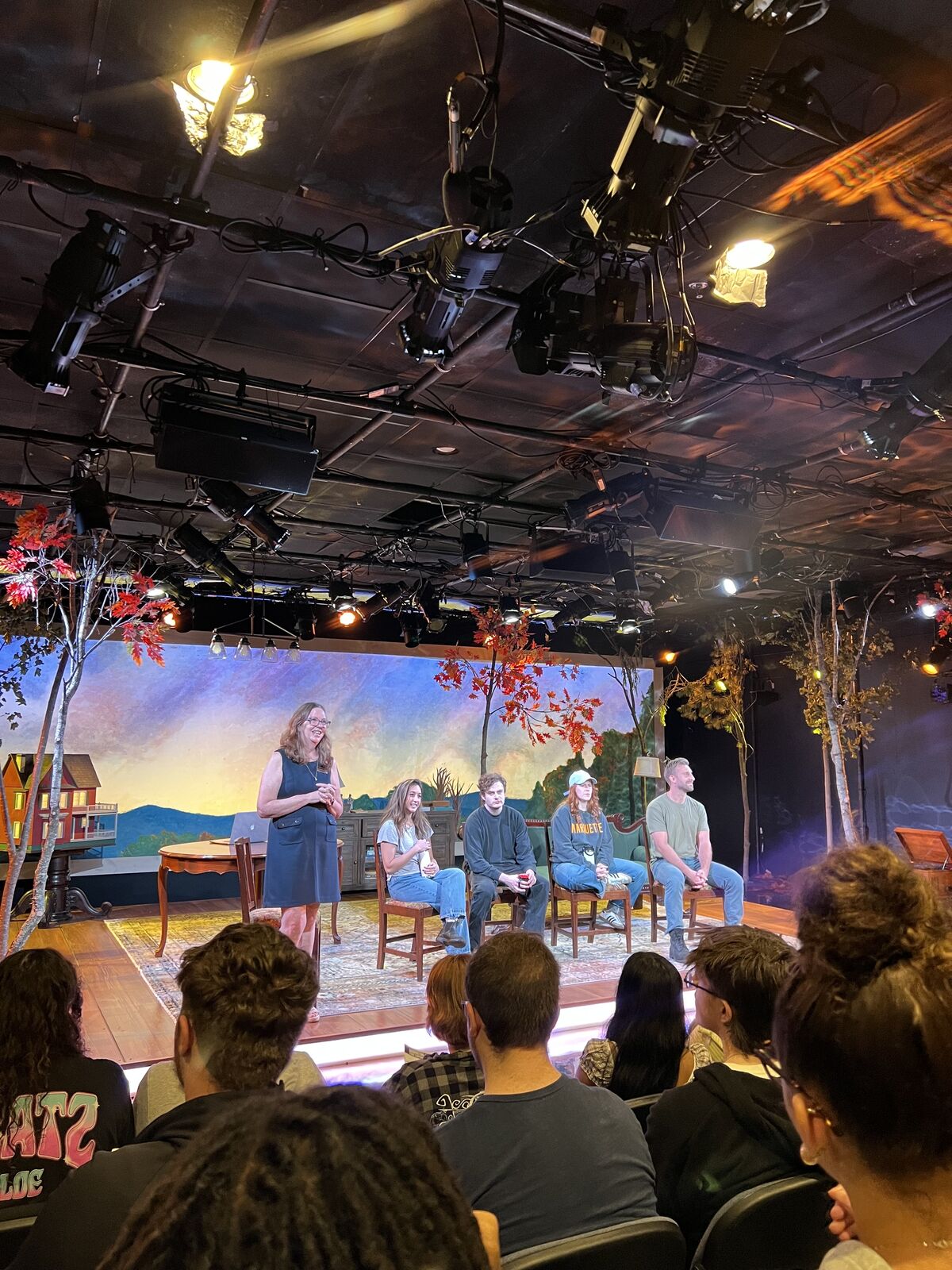 A stage set with a painted backdrop and people seated on chairs, viewed from the audience.