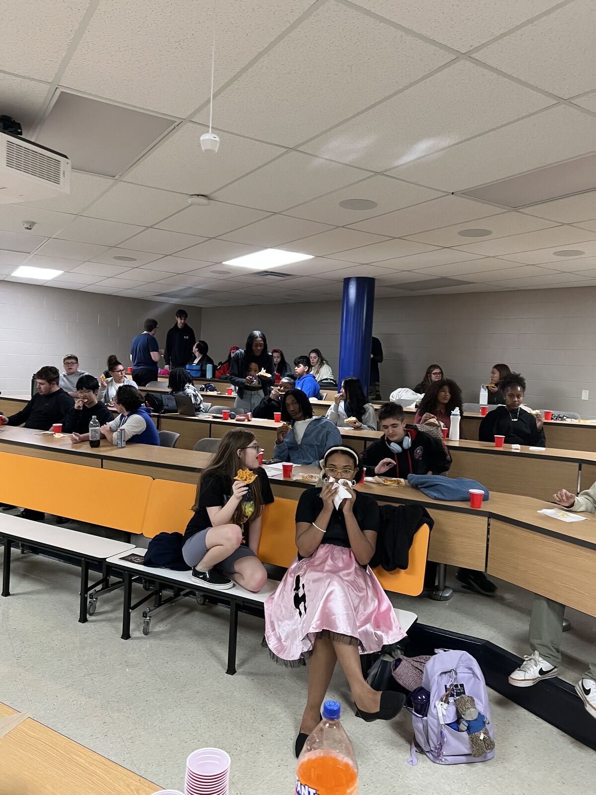 Students eat lunch in a school cafeteria, seated at long tables.