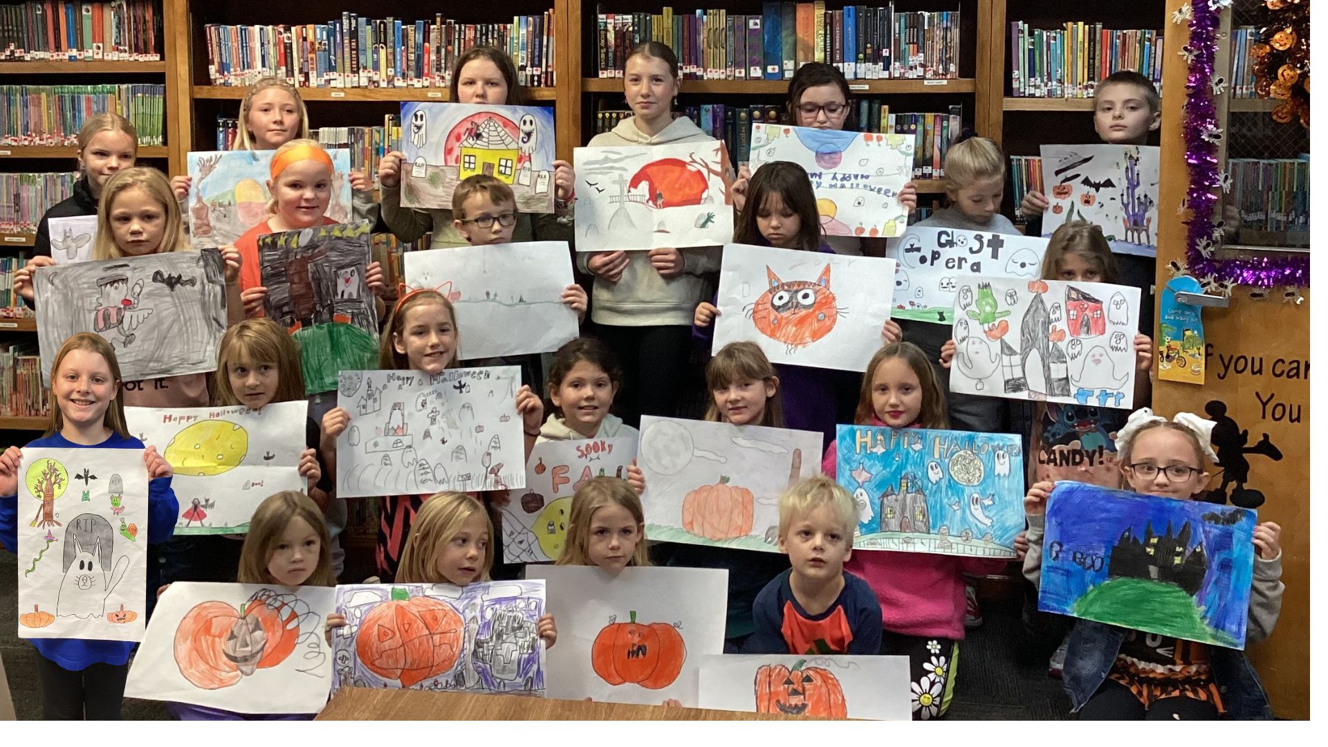 A group of children hold up their Halloween-themed drawings in a library.