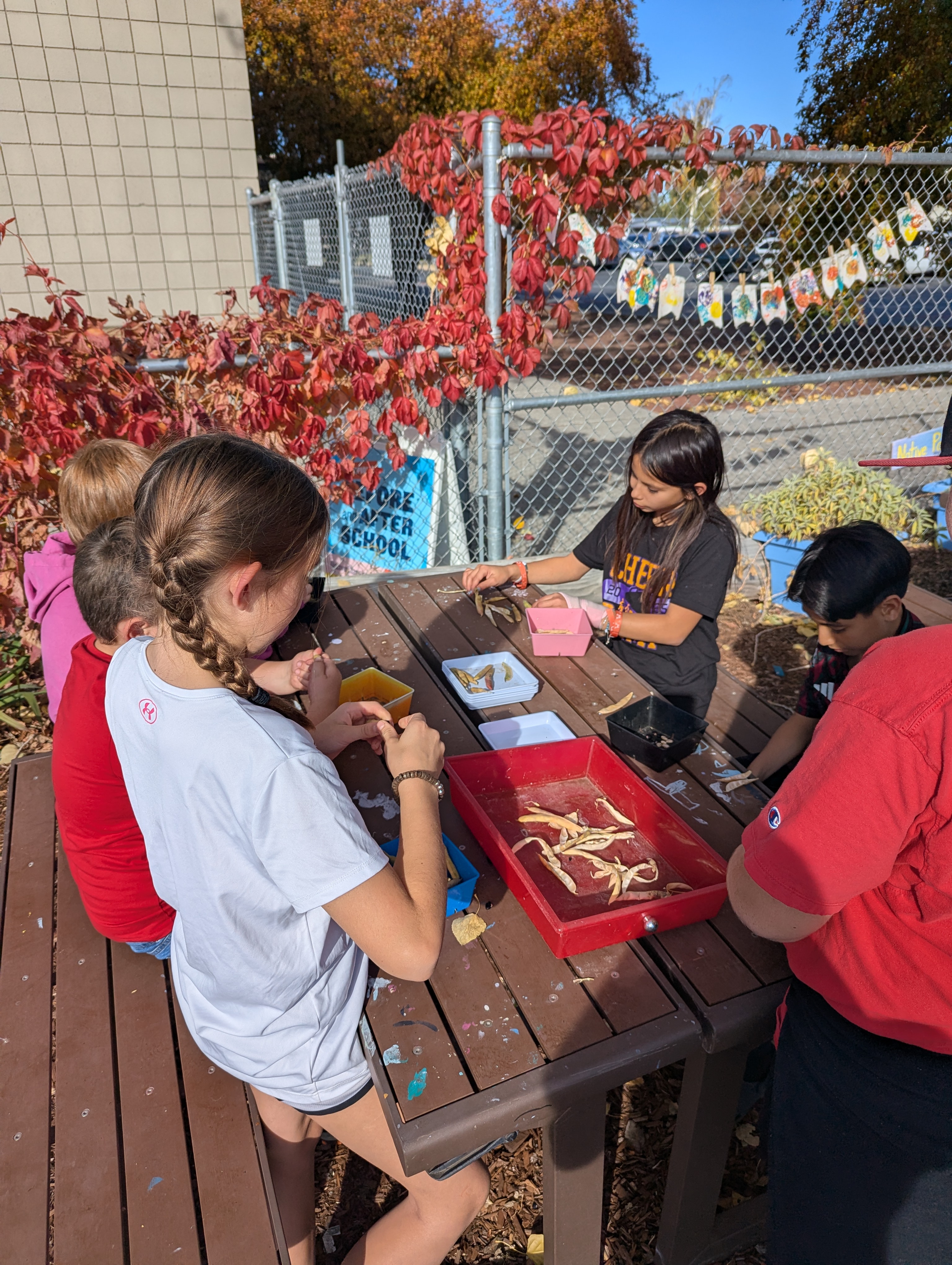 Children gather around a table, examining objects in small containers outdoors.