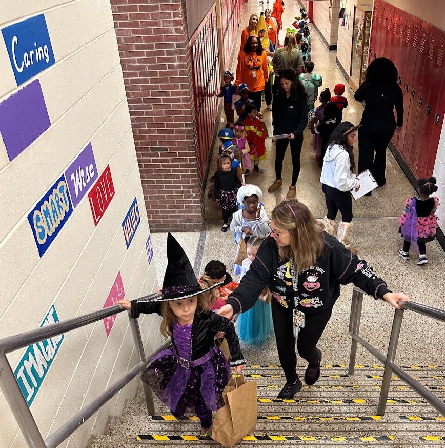 Children in costumes walk down a school hallway, led by adults.