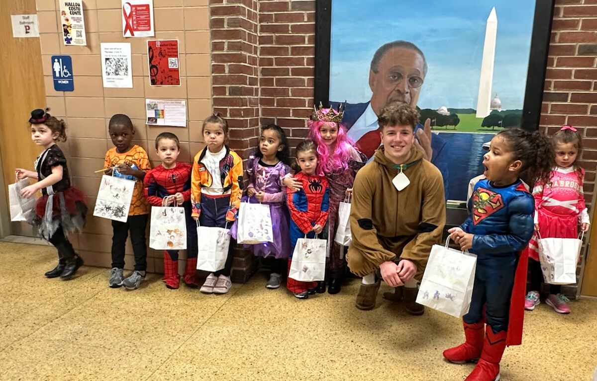 Children in costumes stand in a school hallway, holding treat bags.