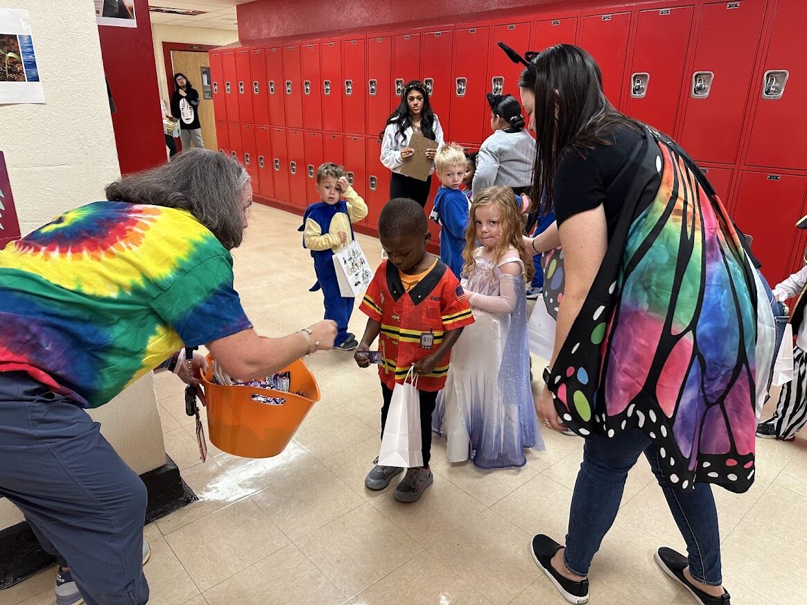 Children in costumes receive treats from an adult in a school hallway.