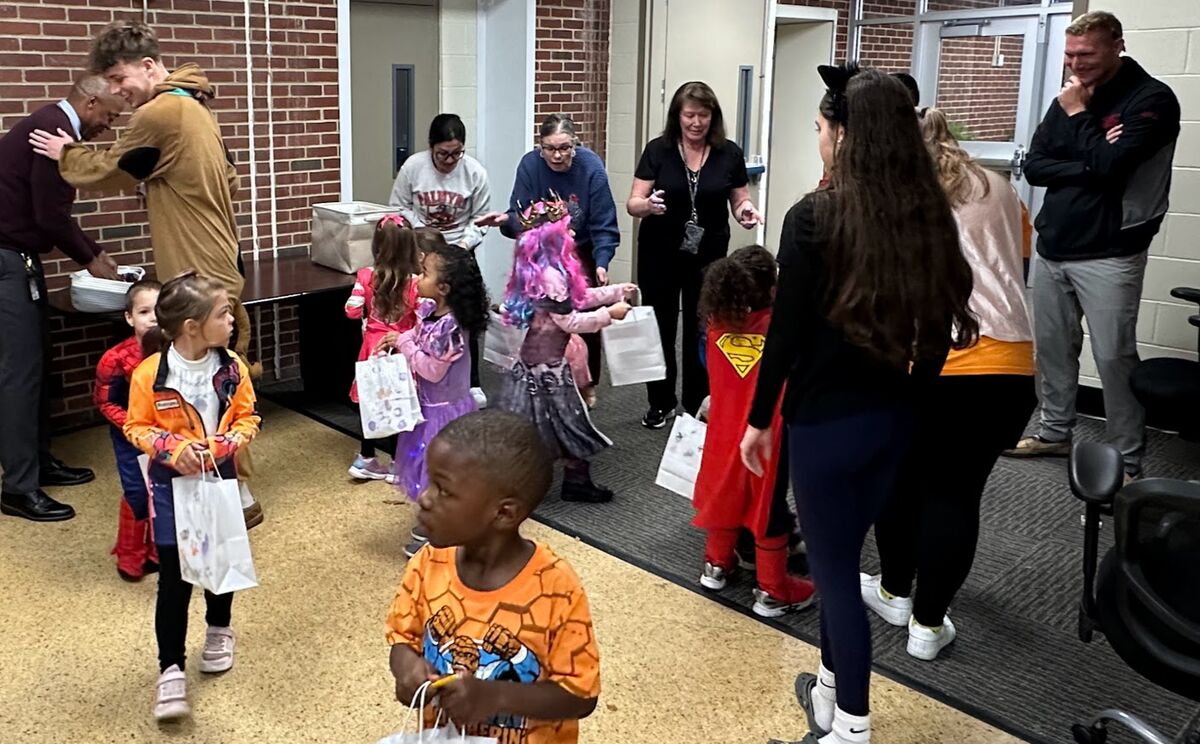 Children in costumes receive treats from adults in a hallway.