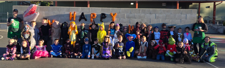 A group of children in Halloween costumes pose in front of a wall with 'HAPPY' balloons.
