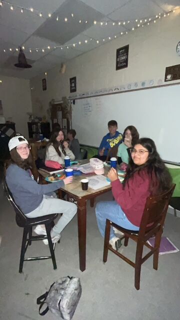 A group of students sit around a table in a classroom, chatting.