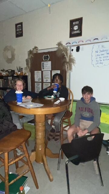 Three students sit around a table in a classroom, enjoying drinks.