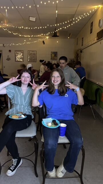 Two young women smile and make peace signs, holding plates of treats.