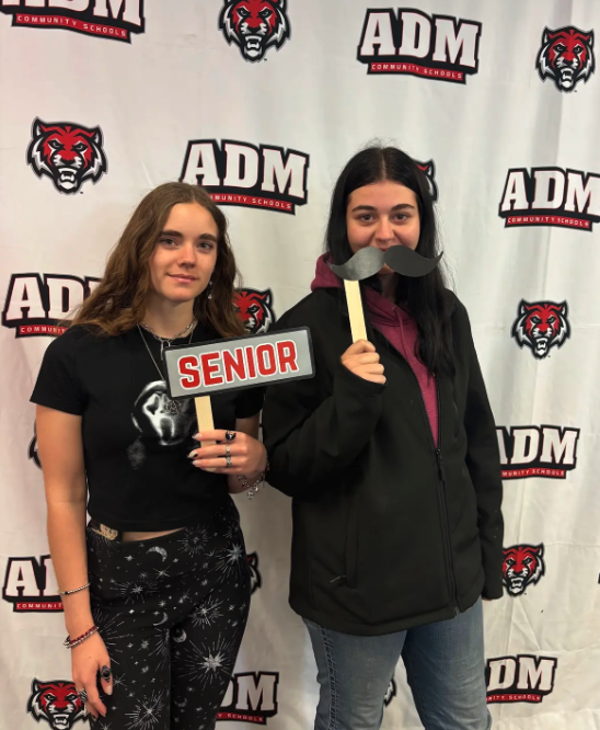Two young women pose for a photo, one holding a 'Senior' sign and the other a mustache prop.