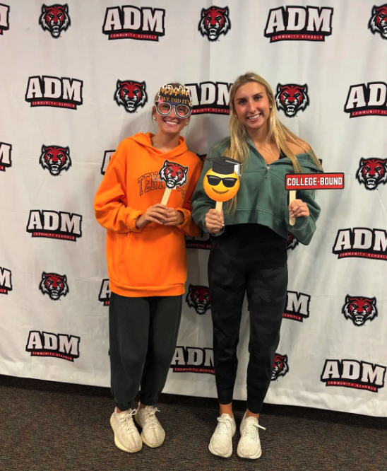 Two young women pose with celebratory props in front of a school banner.