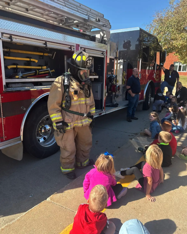 A firefighter in full gear stands near a fire truck, facing a group of children.