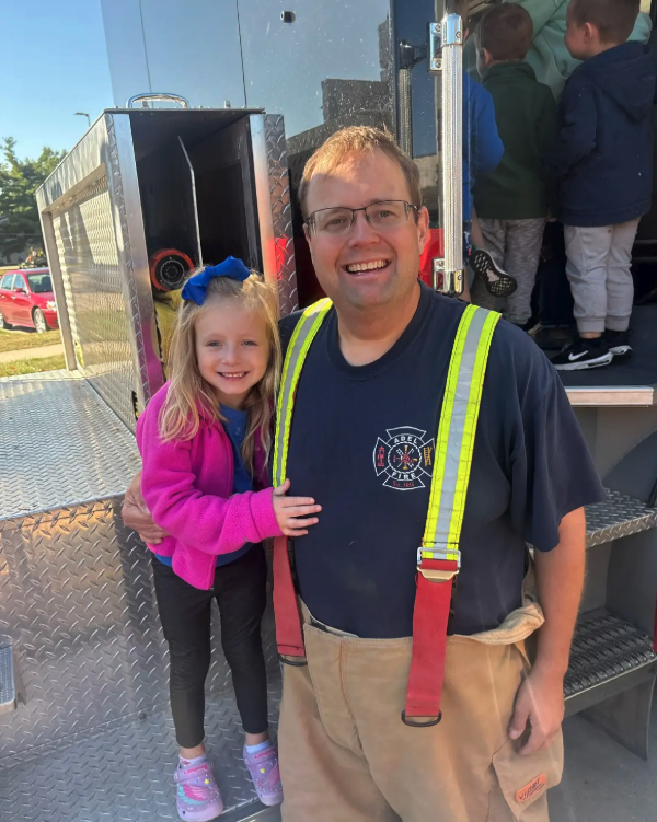 A firefighter and a young girl smile together in front of a fire truck.