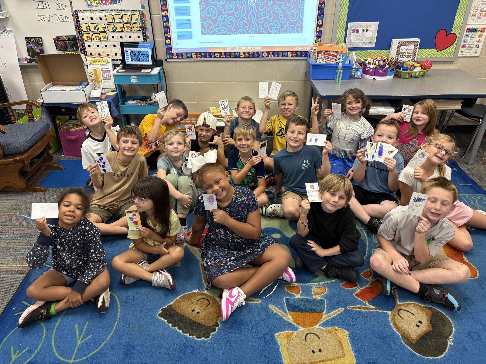 a classroom of 2nd grade students sit clustered on a colorful carpet, smiling while holding up notecards.