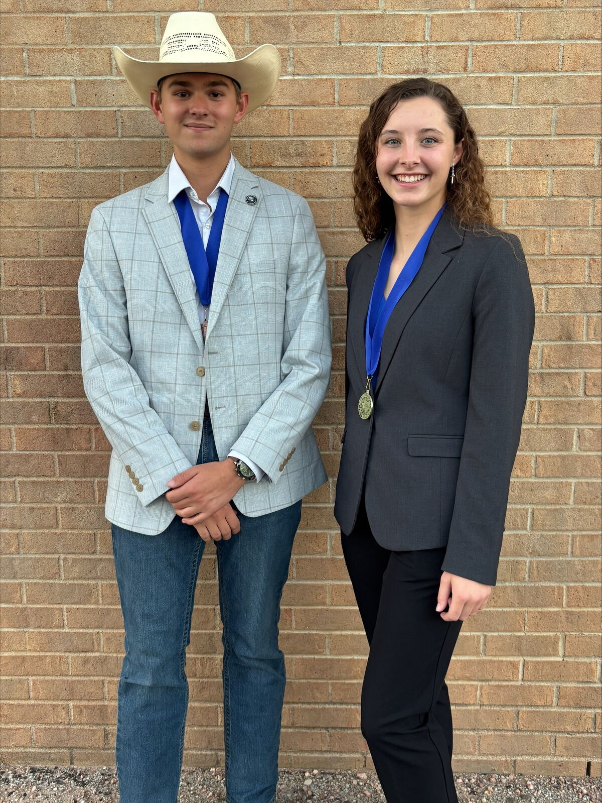 Two young people stand in front of a brick wall, wearing blazers and medals.