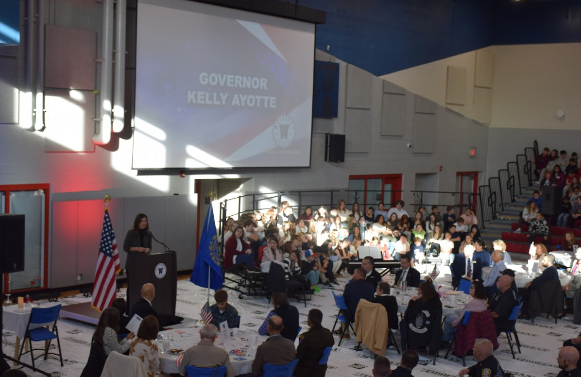 A woman speaks at a podium in front of a large audience, with a screen displaying 'Governor Kelly Ayotte'.