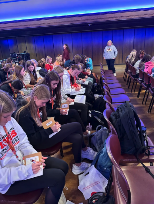 A group of students sit in rows, writing on paper in a large room.