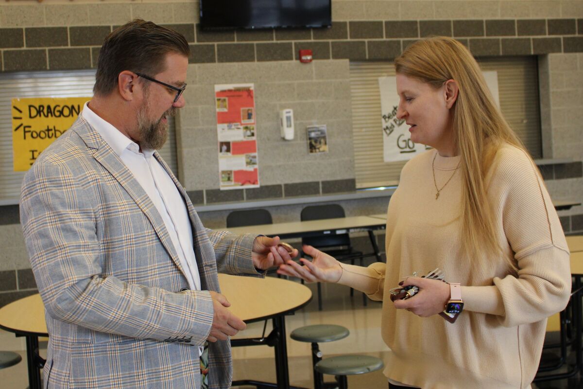 Dr. Robinson hands a commemorative coin to Mrs. Strzyzewski to recognize her military service. They are standing in a school commons.
