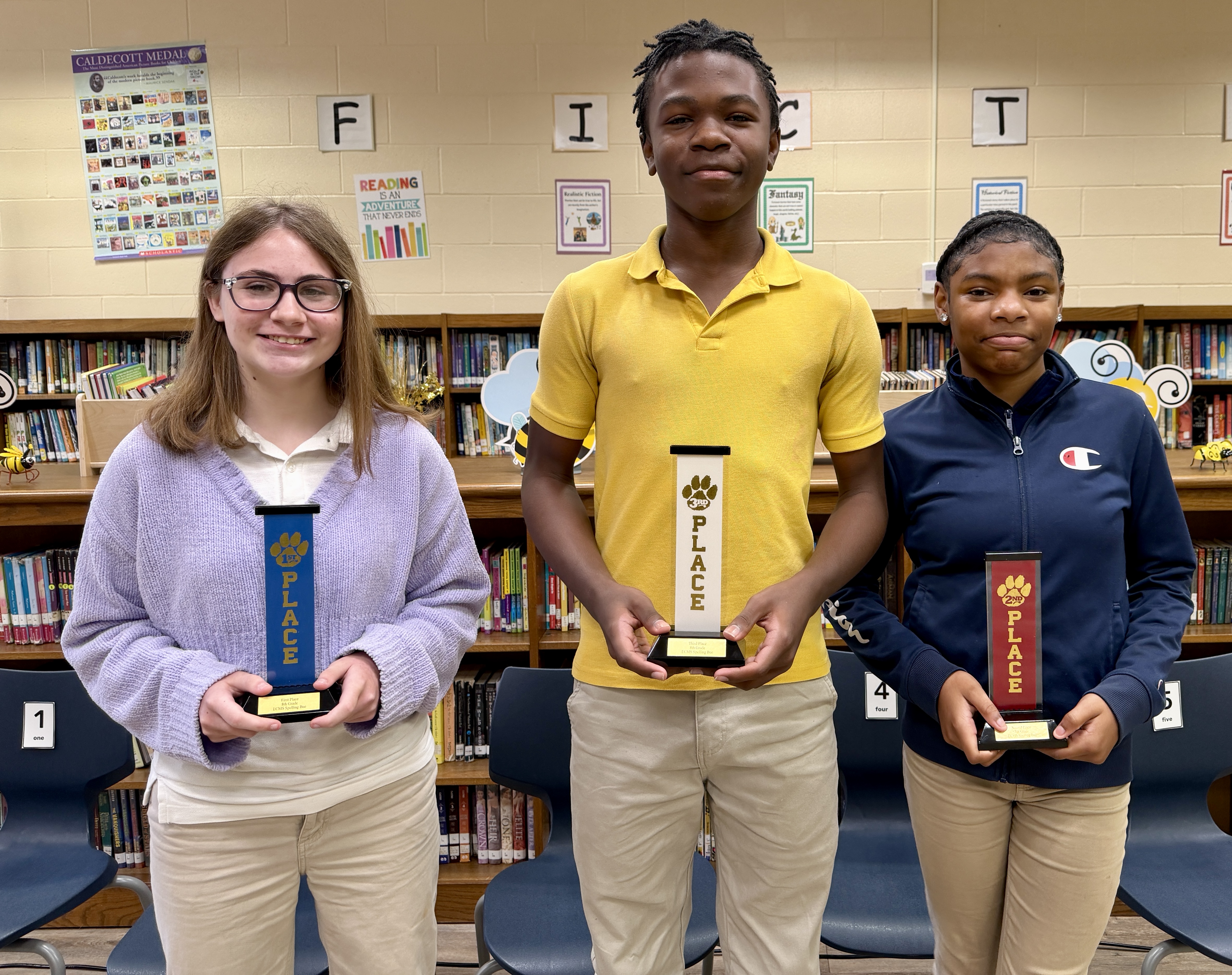 Three students stand proudly, each holding a trophy in a library setting.