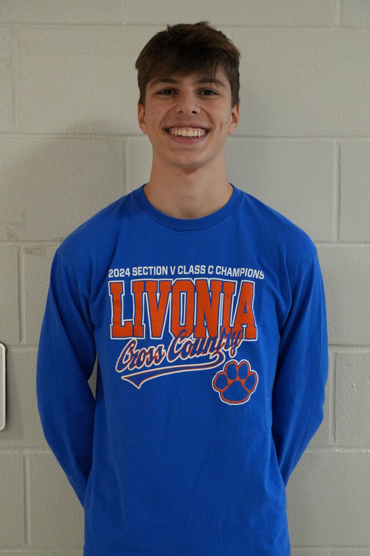 A smiling young man stands in front of a light gray brick wall.