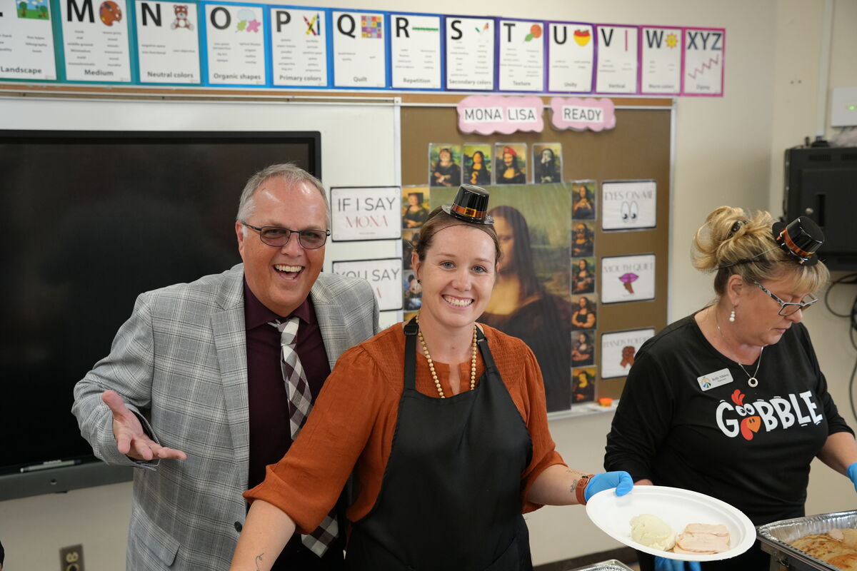 Three people smile in a classroom setting, one wearing a Mona Lisa hat.