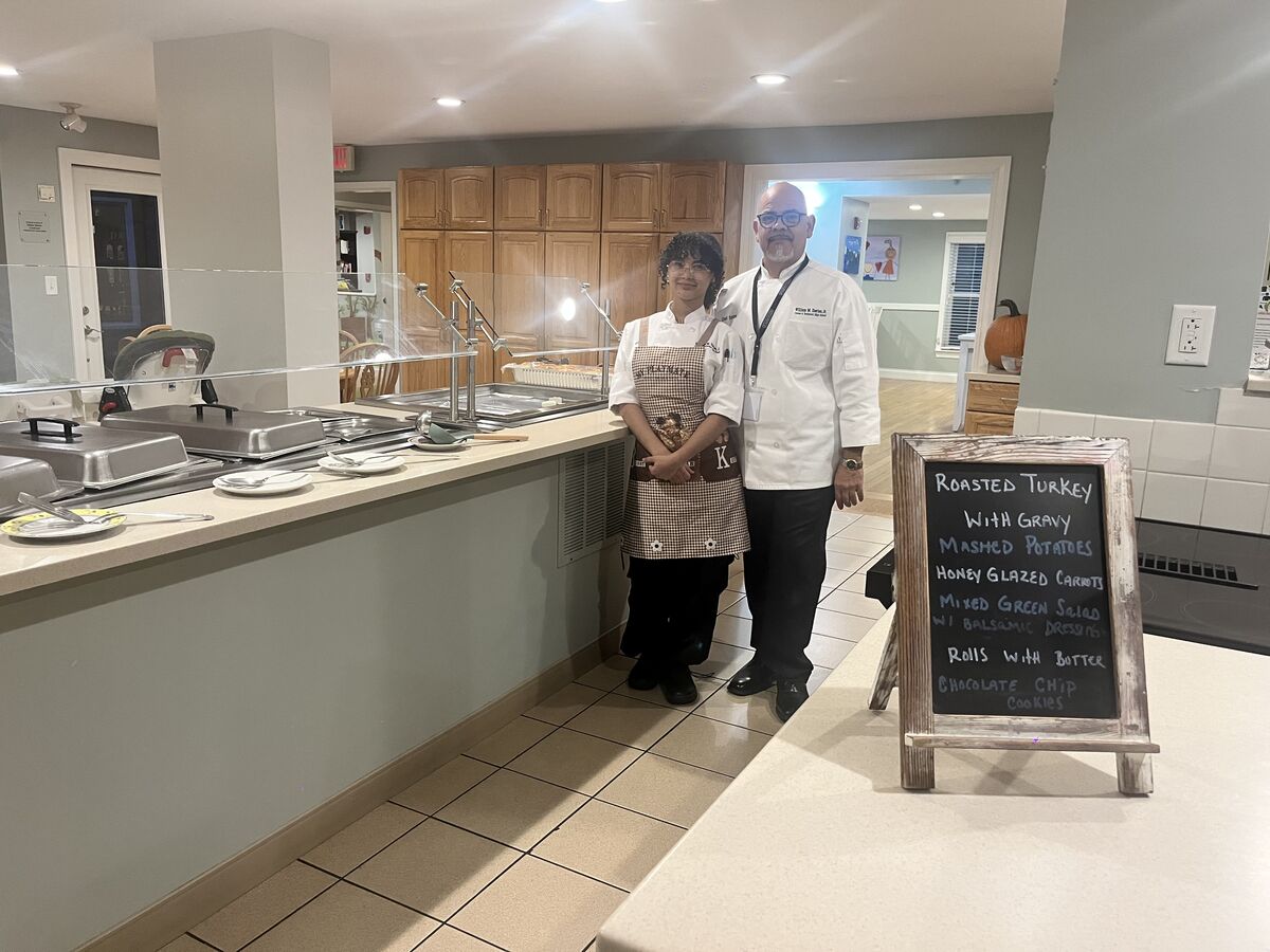 Two chefs stand in a commercial kitchen, posing near a menu board.