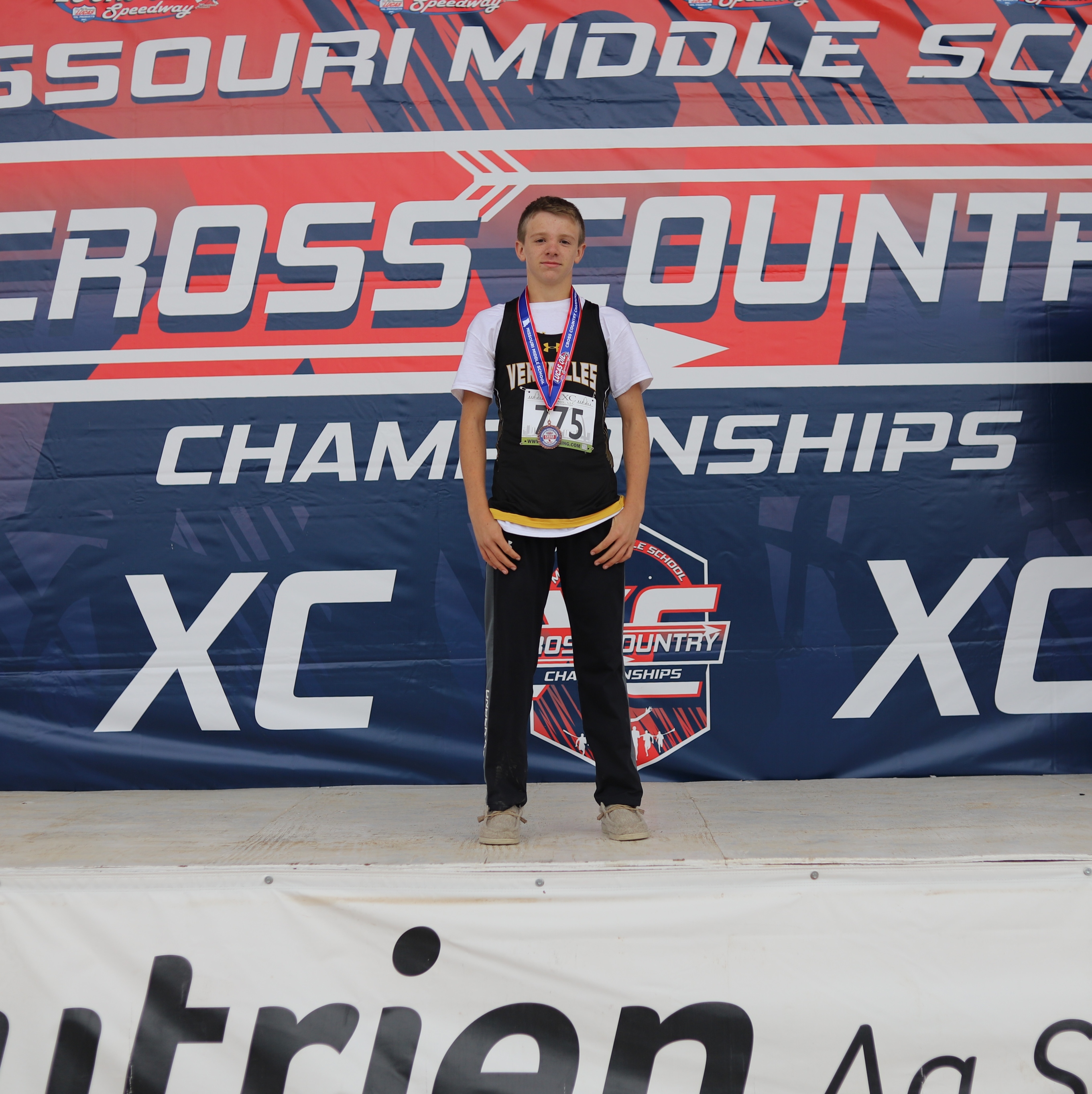 A young athlete stands on a podium, wearing a medal and cross country gear.