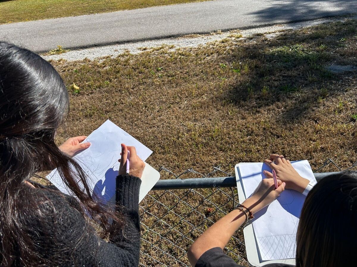 Two people draw on paper outdoors, near a chain-link fence.